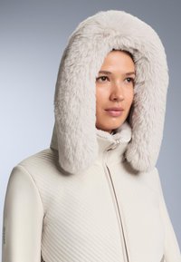 Fehér steppelt dzseki világosszürke plüss műszőrme kapucnival. Sima textúra, első cipzárral és RECCO márkajelzéssel az ujjon.