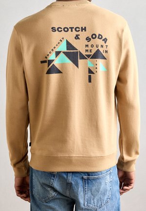 Sweat-shirt beige avec un motif géométrique de montagne noir et turquoise et le texte « Scotch & Soda Mount Me In 1985 » dans le dos, porté avec un jean bleu.