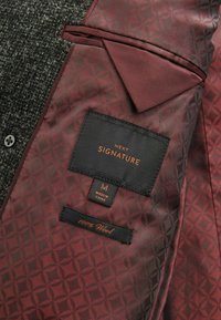 Forro de chaqueta de lana gris oscuro con un patrón geométrico burdeos. Etiquetas: "NEXT SIGNATURE," tamaño "M," y "100% Lana."