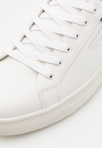 Białe skórzane sneakersy z teksturowaną powierzchnią, płaskimi białymi sznurowadłami, okrągłymi przelotkami i gumową podeszwą z diamentowym wzorem zapewniającym przyczepność.