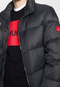 Svart puffjacka med dragkedja och hög krage, prydd med en röd HUGO-lapp. Under jackan bärs en svart sweatshirt med en dj ärv röd grafik.