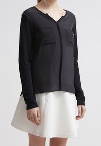 Schwarze Bluse mit langen Ärmeln und V-Ausschnitt, ausgestattet mit zwei vorderen Taschen und einer glatten Textur, kombiniert mit einem strukturierten weißen Rock.