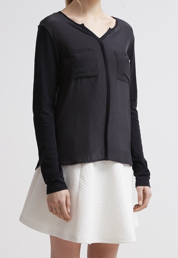Schwarze Bluse mit langen Ärmeln und V-Ausschnitt, ausgestattet mit zwei vorderen Taschen und einer glatten Textur, kombiniert mit einem strukturierten weißen Rock.