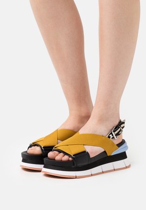 Sandales à plateforme avec un bandeau élastique jaune en croix, une base en cuir noir et une semelle stratifiée multicolore avec un détail de sangle texturée.