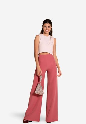 Top court rose clair avec un col rond, associé à un pantalon rose à taille haute et à jambes larges. Elle tient un sac à main en argent pailleté.