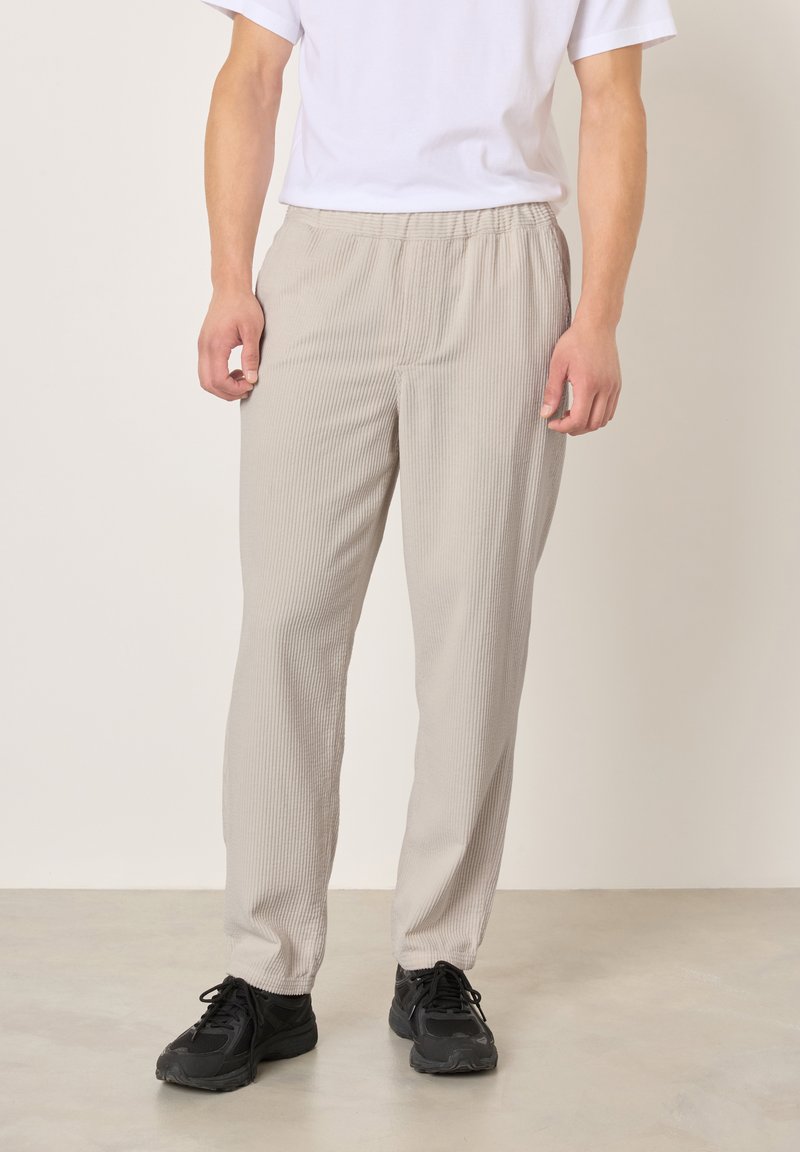 Vyras dėvi smėlio spalvos raukšlėtas kelnes, baltą trumpomis rankovėmis marškinėlį ir juodus sportinius batelius, stovintis ant šviesios grindų dangos priešais paprastą sieną.