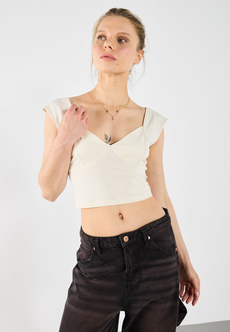 Lys cremefarvet cropped top med kapærmer, der har en tekstureret korpus og en dyb V-udskæring, kombineret med højtaljede mørkeblå denimjeans.