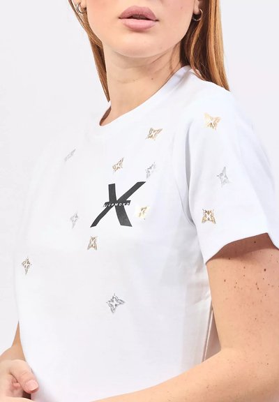 T-shirt bianco in cotone con dettagli metallici a forma di stella in oro e argento, con un logo "X" nero sul davanti. Maniche corte e collo rotondo.