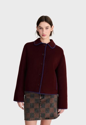 Femme portant une veste bordeaux foncé boutonnée avec un liseré bleu et une jupe à carreaux marron et grise, debout sur un fond clair uni.