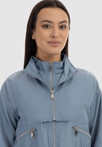 Jachetă albastră deschis, cu fermoar, cu guler înalt, având accesorii din metal argintiu și două buzunare frontale cu fermoar, realizată dintr-un material neted și ușor.