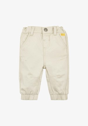 Pantalons en coton beige avec des poignets élastiques, des poches avant et une fermeture à bouton. Présente des détails de couture et une petite étiquette de marque jaune.