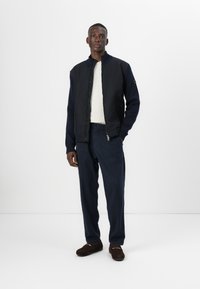 Ναυτικό μπλε μπουφάν bomber, υφασματισμένο, με ριμπ μανσέτες, φορεμένο πάνω από ένα κρεμ πουλόβερ. Ναυτικές παντελόνες και καφέ σουέτ παπούτσια ολοκληρώνουν την εμφάνιση.