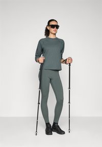 Urheilullinen asu, joka koostuu kuitupintaisesta tummanvihreästä pitkähihaisesta paidasta ja siihen sopivista legginsseistä, asustettuna mustilla vaellussauvoilla ja kengillä.