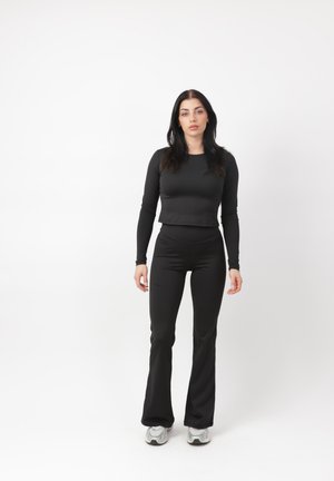 Kobieta o długich ciemnych włosach, ubrana w dopasowaną czarną bluzkę z długim rękawem i czarne spodnie, stojąca na jednolitym białym tle.