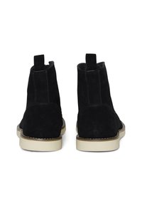 Botas de tobillo de ante negro con lengüetas, upper texturizado y suelas de goma de color claro en contraste. Presenta costuras a lo largo de los bordes de la suela.