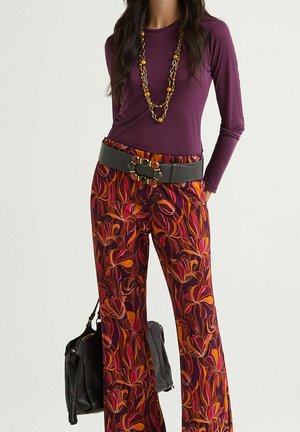 Maglia viola a maniche lunghe con design aderente, abbinata a pantaloni a gamba larga con motivo floreale arancione e rosa. Cintura nera con dettagli decorativi in metallo.