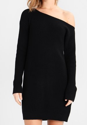 Kobieta w czarnej, dzianinowej sukience swetrowej z dekoltem opadającym na ramię odsłaniającym lewe ramię.