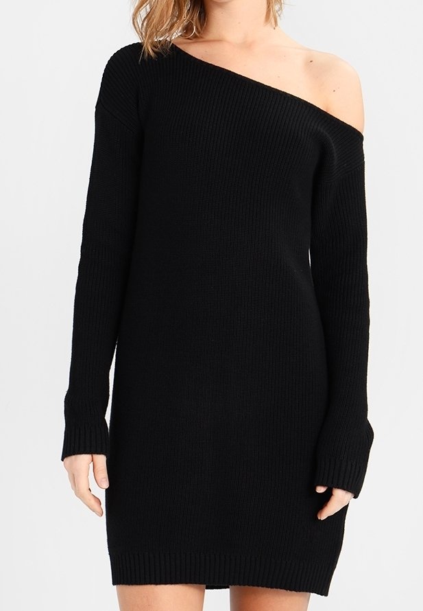 Femme portant une robe pull noire en tricot avec un décolleté épaules dénudées exposant l'épaule gauche.