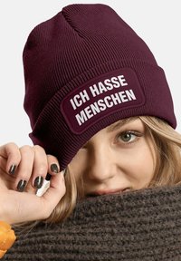 Burgundy knit beanie featuring a wide patch with white text "ICH HASSE MENSCHEN." Close-up view with a textured dark scarf in the background.