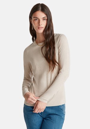 Junge Frau mit langen dunklen Haaren trägt ein beiges Langarmoberteil, blaue Jeans, eine goldene Kettenhalskette und goldene Ohrstecker vor einem weißen Hintergrund.