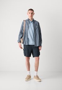 Tenue décontractée comprenant une chemise à carreaux bleue, un polo bleu clair, un short cargo sombre et des baskets beiges, le tout complété par un sac à épaule beige.