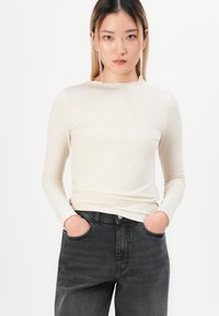 Ung kvinne med langt hår iført en tettsittende kremfarget langermet topp og løse høytlivs mørkegrå jeans, hendene i lommene, nøytral bakgrunn.