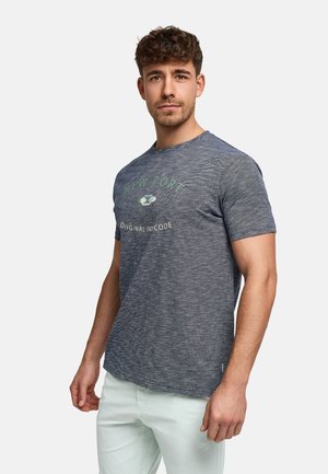 Junger Mann mit lockigem braunem Haar, der ein blaues New Port Original Code T-Shirt und helle Hosen trägt, vor einem einfarbigen Hintergrund steht.