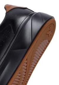 Primo piano di una scarpa in pelle nera con una suola intermedia nera spessa e una suola esterna in gomma marrone testurizzata con un piccolo logo impressa.