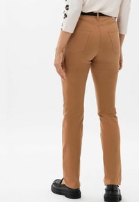 Femme portant un pantalon beige ajusté, une ceinture noire, un haut blanc avec des manches à motifs, et des chaussures compensées noires, debout devant un fond uni.