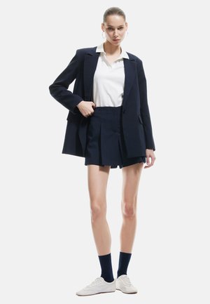 Marineblazer met gestructureerde schouders, witte poloshirt, geplooide marineblauwe shorts en witte sneakers. Het model staat voor een neutrale achtergrond.