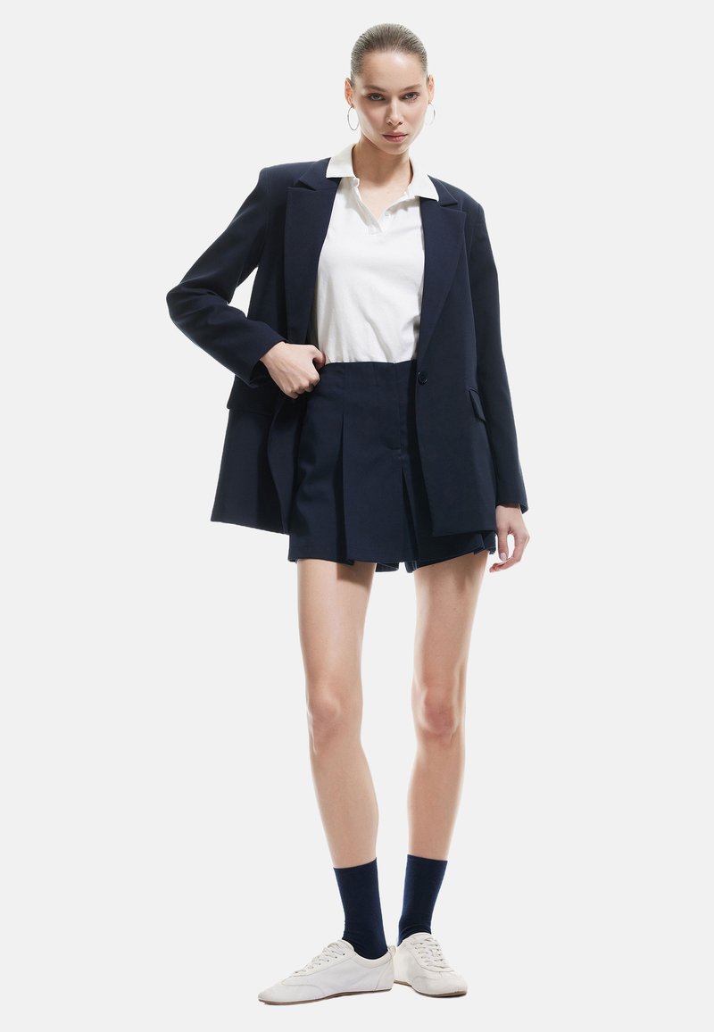 Laivastonsininen bleiseri, jossa on jämäkät olkapäät, valkoinen poolopaita, taitettu laivastonsininen shortsit ja valkoiset tennarit. Malli seisoo neutraalissa taustassa.