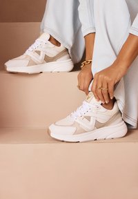 White and beige athletic sneakers with mesh and suede textures, featuring a chunky white sole and a layered design. Accented with laces.