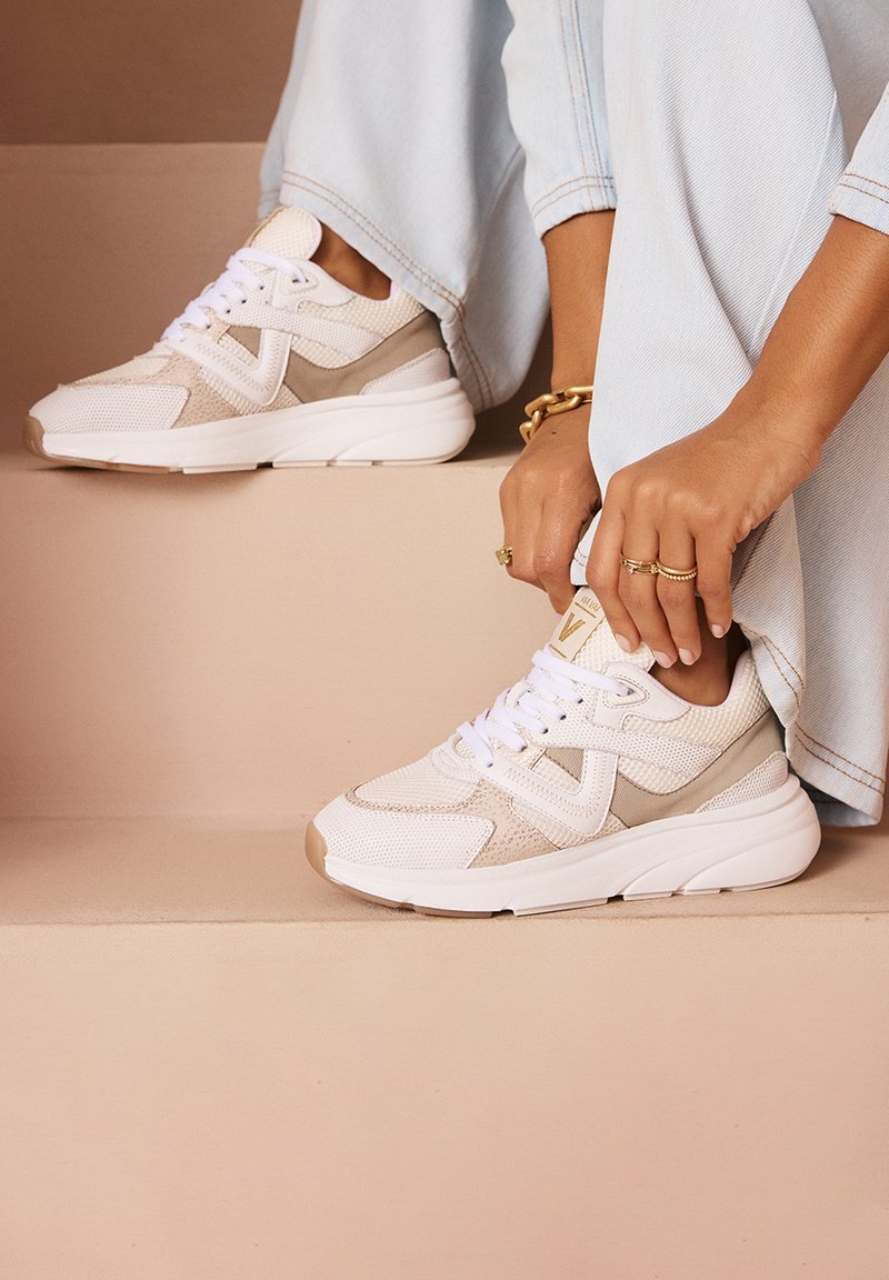 White and beige athletic sneakers with mesh and suede textures, featuring a chunky white sole and a layered design. Accented with laces.