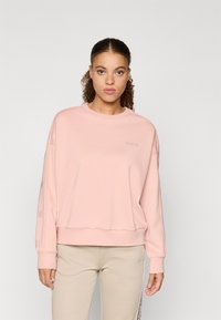 Világos rózsaszín pulóver ribbelt mandzsettával, kerek nyakkal, és dombornyomott logóval a mellkason. Puha anyag és laza szabás.