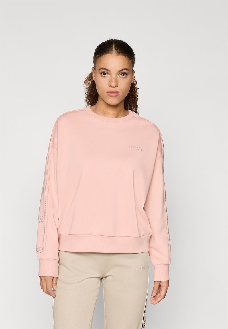 Világos rózsaszín pulóver ribbelt mandzsettával, kerek nyakkal, és dombornyomott logóval a mellkason. Puha anyag és laza szabás.