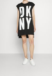 Robe t-shirt noire avec un grand lettrage blanc « DKNY », col rond et manches courtes, portée avec des baskets blanches.