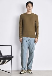 Brauner, gerippter Strickpullover, getragen mit hellblauen Jeans und mehrfarbigen Sneakers. Das Model steht in einem minimalistischen Ambiente neben einem schwarzen Stuhl.