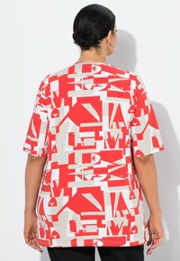 Červené a béžové oversized tričko s geometrickým vzorem písmen, krátkými rukávy a kulatým výstřihem, vyrobené z jemné a hladké látky.