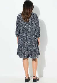 Schwarz-weißes Kleid mit floralem Muster, drei Viertelärmel und gestuften Rock. Glattes Material, knielang, getragen mit schwarzen Sandalen.