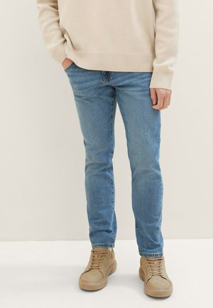 Hellblaue Jeans im Slim-Fit kombiniert mit beigen Sneakers. Die Textur der Jeans ist leicht ausgewaschen und weist subtile Details auf.