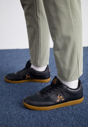 Zapatillas negras con malla texturizada y cuero liso, con un logotipo marrón, suela de goma y calcetines blancos, combinadas con pantalones verde claro.