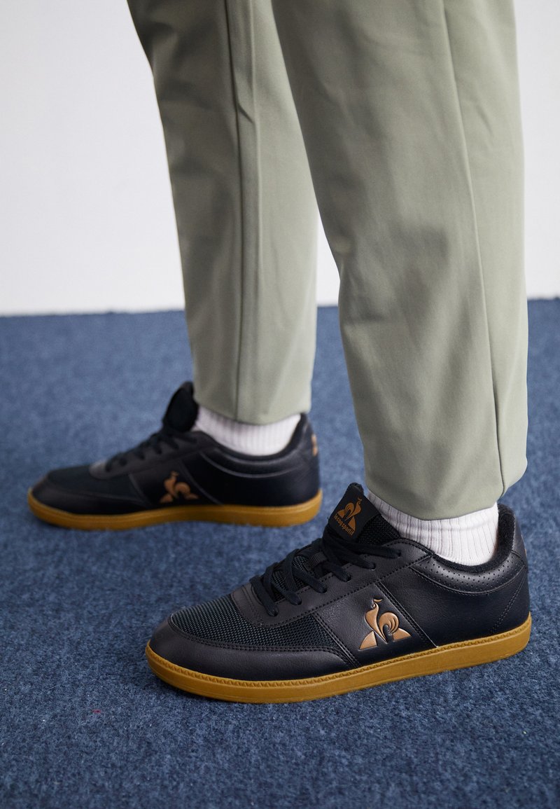 Des baskets noires en maille texturée et cuir lisse, arborant un logo marron, une semelle en caoutchouc beige, et des chaussettes blanches, associées à un pantalon vert clair.
