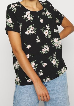 Femme portant un haut noir à manches courtes avec un imprimé floral rose et blanc et un jean en denim bleu clair, debout avec un bras plié.