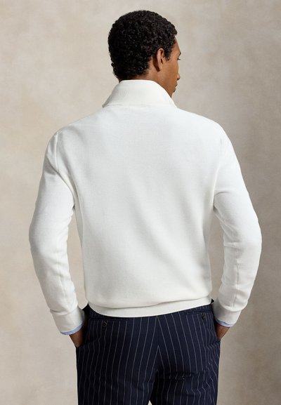 Homme de dos portant un pull texturé blanc à manches longues et un pantalon bleu marine à fines rayures, les mains dans les poches.