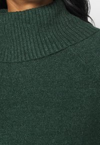 Gros plan sur une personne portant un pull à col roulé côtelé vert foncé avec une texture de tricot visible et une couture à l'épaule.