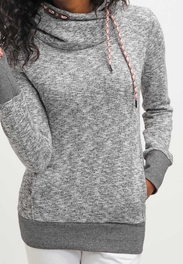 Femme portant un sweat à capuche à manches longues gris chiné avec un col boule et des cordons tressés, assorti à un pantalon blanc.