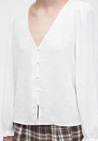 La femme porte un chemisier blanc texturé à manches longues avec des boutons recouverts de tissu et un col en V, associé à une jupe à carreaux marron et blanche.