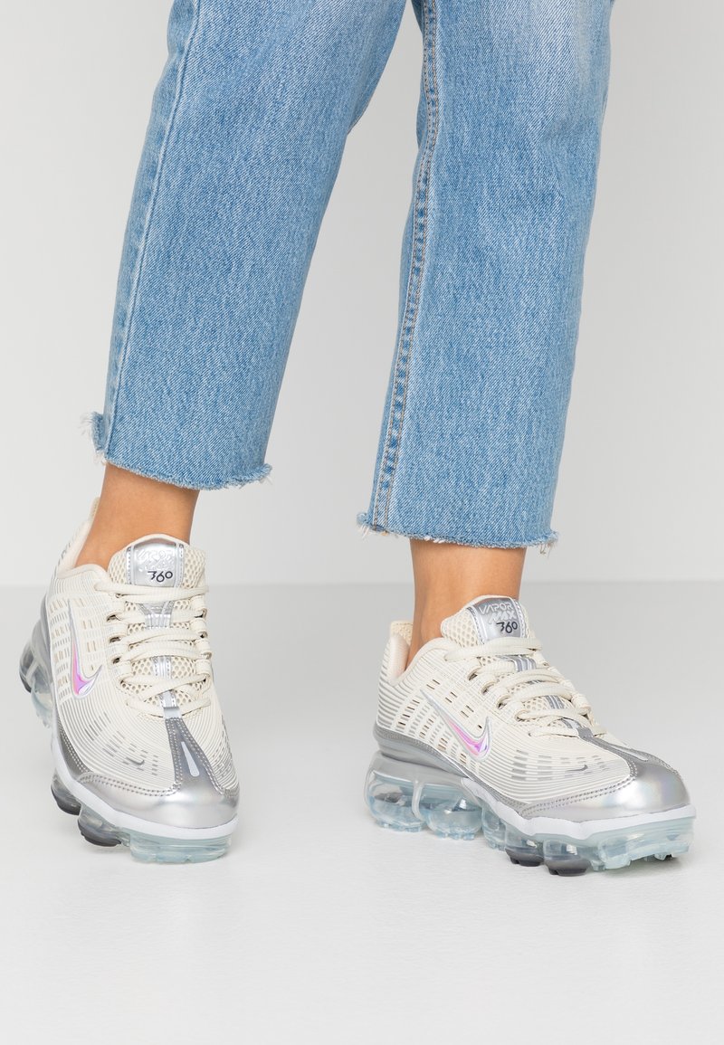 Des pieds portant des baskets Nike Vapormax 360 blanches et argentées avec des semelles translucides et un jean bleu clair aux ourlets effilochés.