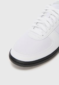 Sneaker branco de malha com uma textura suave, língua acolchoada, atacadores brancos planos e uma sola de borracha preta ao longo das bordas.