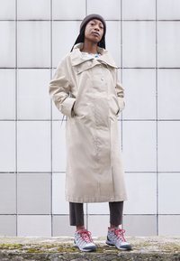 Beige vattentålig trenchcoat med stor krage och knappar, tillsammans med bruna byxor och grå vandringsskor med röda snören. Brunt stickat tygmössa bärs.